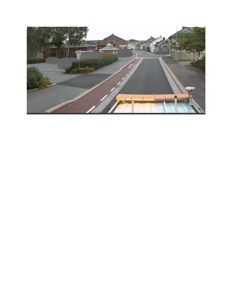 Fotografi tatt fra en kjørende bil (sannsynligvis en lastebil eller buss) som viser en gate i et boligområde med hus og parkeringsplasser.