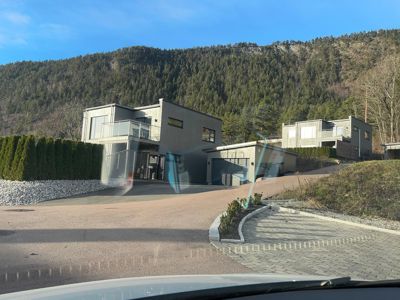 Fotografi av en moderne bolig sett fra en vei, med en skogkledde bakke i bakgrunnen.