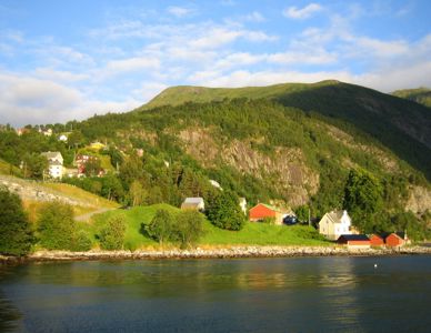 Fotografi av en kystbygd med hus på en grønn, skogkledt bakke ved vannet.