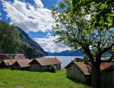 Fotografi av en bygning eller flere småbygninger (trolltun) ved en vannflate, med fjell i bakgrunnen.