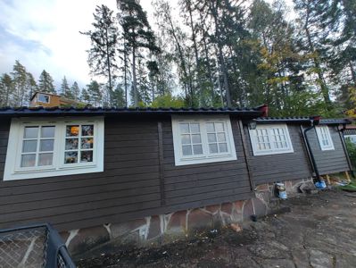 Fotografi av en mørk, trepanelt bygning med hvite vinduer og steinfundament, sett fra siden med skog i bakgrunnen.