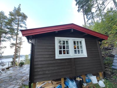 Fotografi av en ferdig eller nesten ferdig bygning (en liten hytte eller uthus) sett fra siden. Bildet viser fasaden med mørke paneler, hvite vinduer og et rødt tak, samt omgivelsene med trær, vann og en steinlagt terrasse.