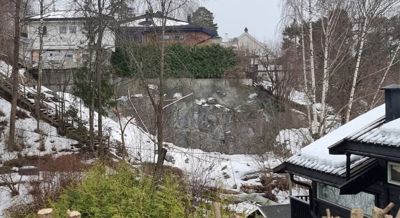 Fotografi av en boligområde med snødekke, trær og en steil skrent eller klippvegg.