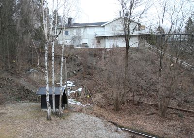 Fotografi av en tomt med en eksisterende hvit villa på bakken og en liten hytte i forgrunnen.