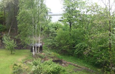 Fotografi av en grønn, trærrik hage eller tomt med en liten hytte og en del av en større bygning i bakgrunnen.