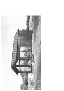 Sort-hvitt fotografi av en liten, moderne hytte eller uthus sett fra siden, med en hage i bakgrunnen.