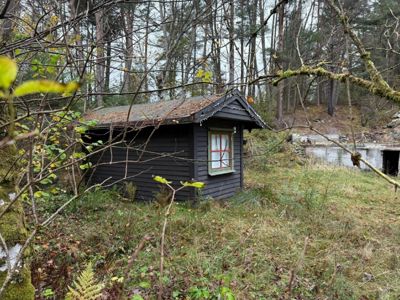 Fotografi av en liten, mørk hytte eller uthus i skogen, sett gjennom grener.