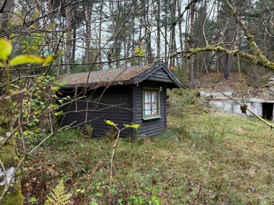 Fotografi av en liten, mørk hytte eller uthus i en skogstomte med et vannløp i bakgrunnen.