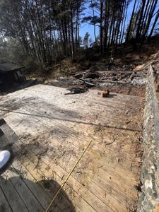 Fotografi av en byggeplass eller en delvis nedrevet bygning i en skog. Bildet viser en utvidet betongplate eller fundament, rester av en vegg, og en tilstøtende hytte i bakgrunnen.
