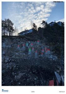Fotografi av en steil fjellside med en utgravet eller utskåret del, sannsynligvis en bergvegg eller en del av en grunnmur. Det er flere hus synlige på toppen av bakken. Bildet har markeringer (røde og grønne sirkler og trekanter) som antyder punkter for boring, uttag eller måling.