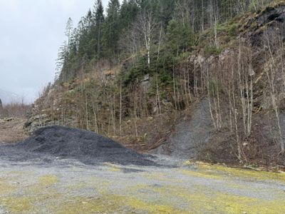 Fotografi av et utemiljø med en stor haug med mørkt materiale (kanskje grus eller asfalt) foran en skråning med trær og stein.