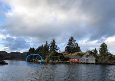 Fotografi av en kystlinje med hus, trær og fjell i bakgrunnen. En blå sirkel er tegnet inn over en del av landskapet.