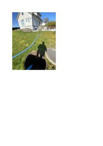 Fotografi av en eksisterende hvit villa sett fra bakgården. Bildet viser en blå slange som ligger på gresset og fører ned i et åpent manuellom (sjekkbrønn) i forgrunnen. Det er også synlig en skygge av fotografen.