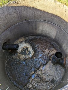 Fotografi av en utvendig avløpsbrønn eller septiktank med synlig slammass og rørforbindelser.