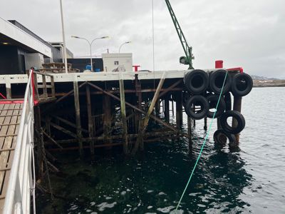 Fotografi av en kai eller pille med tyrebeskyttelse, en bygning i bakgrunnen og vann.