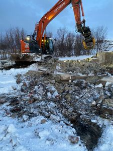 Fotografi av en byggeplass der en gaffeltruck (Hitachi) jobber med å rive eller flytte murverk i snø. Bildet viser en eksisterende eller delvis riven bygning.