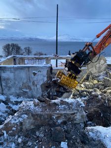 Fotografi av en bygge- eller riveplass med en gaffeltruck som demonterer en betongkonstruksjon, med snø og fjell i bakgrunnen.