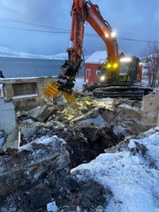 Fotografi av en bygge- eller riveplass med en gaffeltruck som bryter ned betongfundament i snø. I bakgrunnen er det en rød bygning og vann.