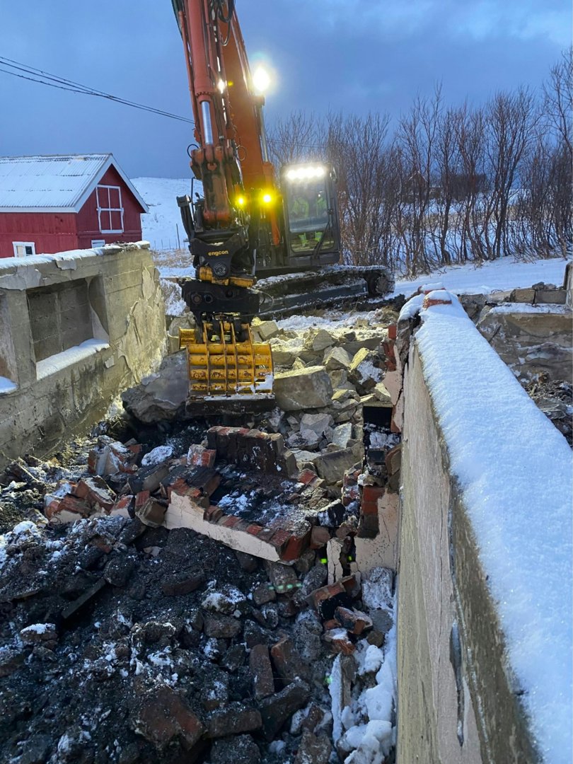 Fotografi av en byggeplass eller riveplass der en gaffeltruck (graver) jobber med å bryte ned en mur. Det er snø på bakken og på en del av muren, og det er en rød bygning i bakgrunnen.