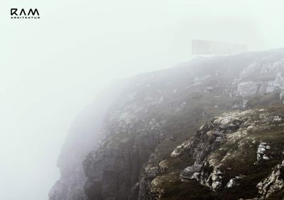 Fotografi av en steil fjellside med tåke og en liten, moderne bygning på toppen.