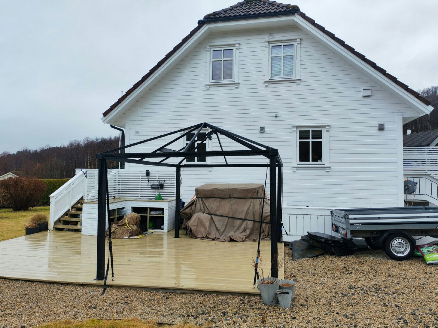 Fotografi av en hvit villa sett fra bakken, med en nybygd terrasse, en svart metallkonstruksjon (gazebo) og en tilhenger foran.