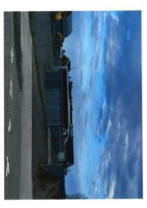 Fotografi av en bygning sett fra siden, med en vei i forgrunnen og en skyet himmel.