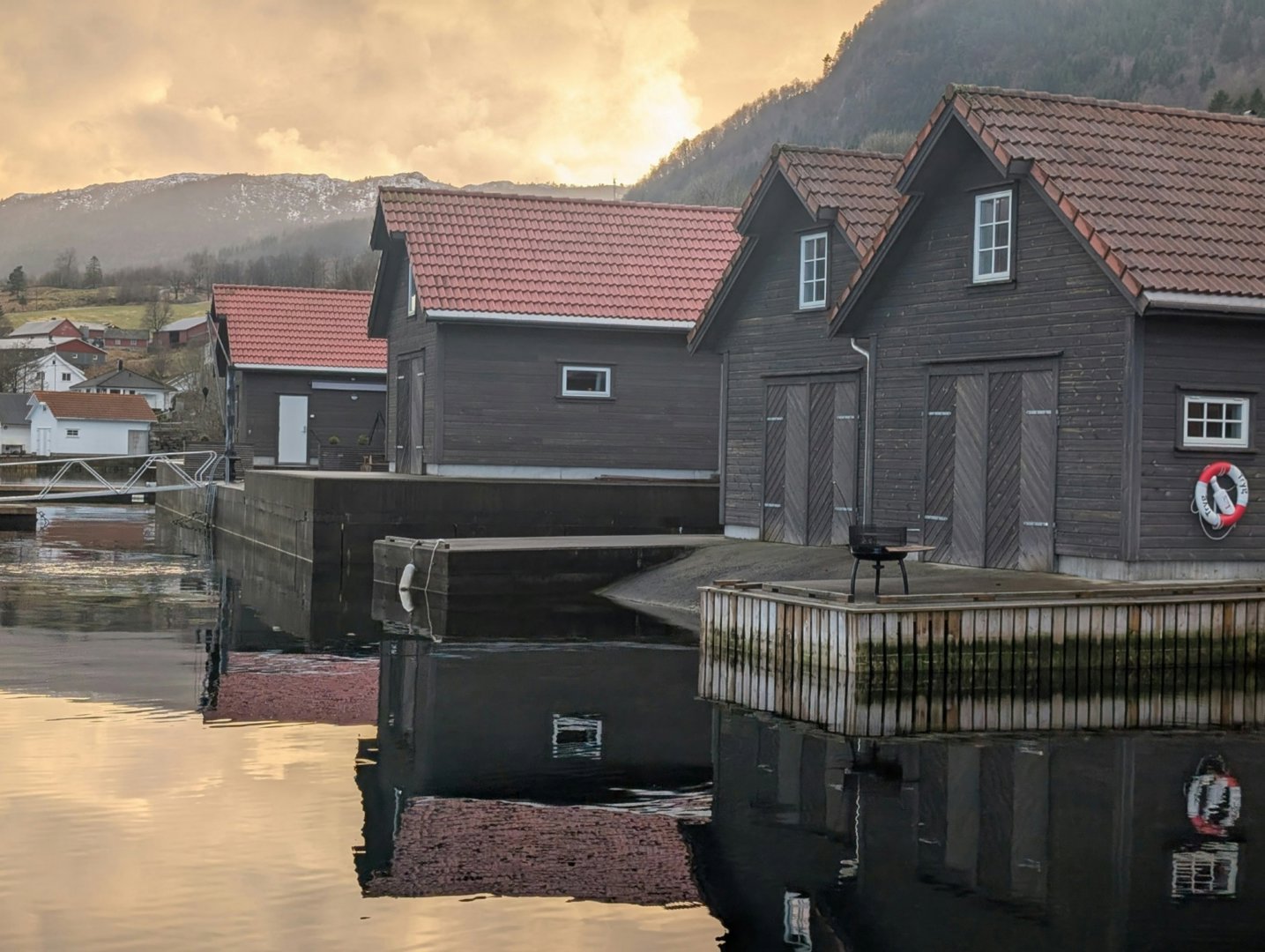 Fotografi av en rekke mørke trehus med røde tak langs en vannkant, med fjell i bakgrunnen.