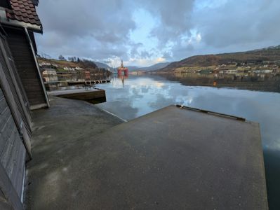 Fotografi av en bygning ved vannkanten med utsikt over et vann, en oljeplattform i bakgrunnen og en by i fjellet.