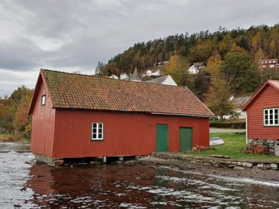 Fotografi av en rød, langbygget bygning (sannsynligvis en uthus eller båthus) som står på stolper ved vannkanten. I bakgrunnen ser man en skogkledt bakke med flere andre hus.