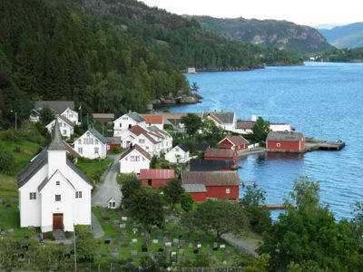 Fotografi av et landsbyområde langs en kyst med en kirke, gravplass og flere hus.
