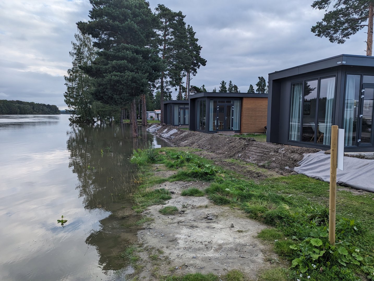 Fotografi av moderne hytter eller boliger plassert langs en vannkant, med natur og terreng i forgrunnen.