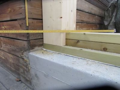 Fotografi av en konstruksjonsdetalj i en bygning, som viser en møte mellom en betongfundament, en trekonstruksjon og en målebånd.