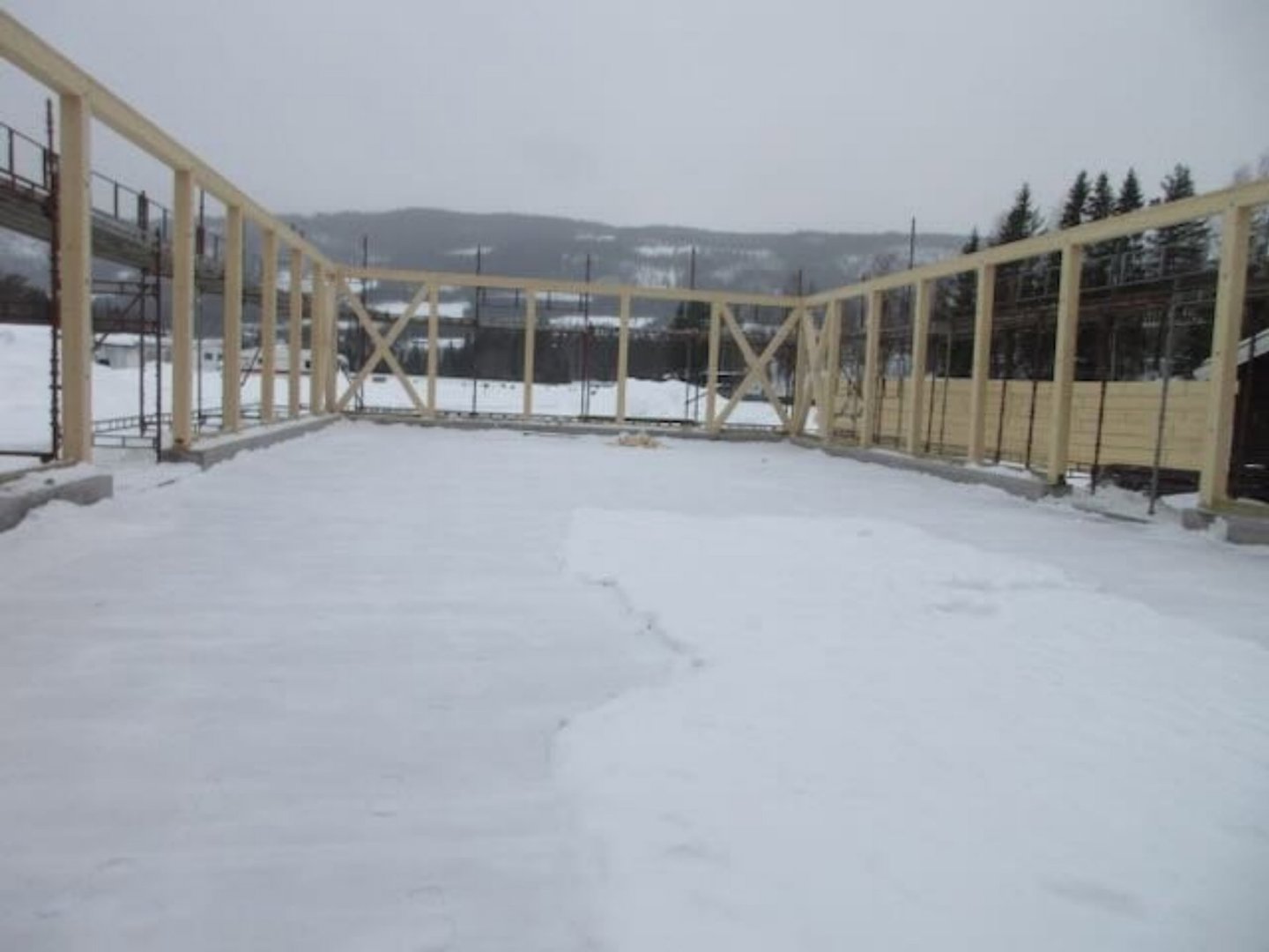 Fotografi av en byggeplass i vinterforhold. Bildet viser et nytt hus under oppføring med oppstilt tømmerkonstruksjon (rammeverk) for vegger og tak. Det er mye snø på bakken og taket, og det er et fjellandskap i bakgrunnen.