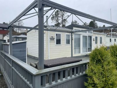 Fotografi av en hvit sommerhytte med en grå metallkonstruksjon (kanskje en veranda eller takkonstruksjon) foran. Bildet viser bygningens fasade, vinduer, en terrasse med møbler og omgivelsene.