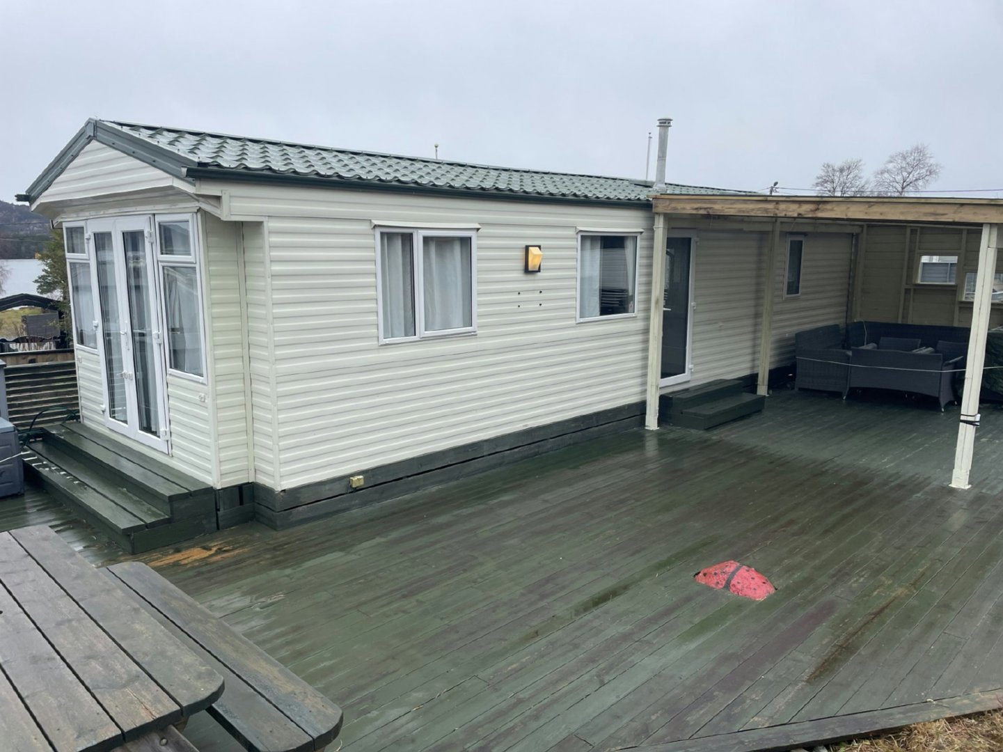 Fotografi av en ferdigbygd hytte eller mobilheim sett fra siden, med en stor terrasse foran.
