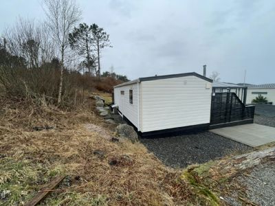 Fotografi av en hvit, modulær hytte eller containerbolig plassert på en skråning. Bildet viser fasaden, en svart hekk, en betongplate og omgivelsene med trær og grus.
