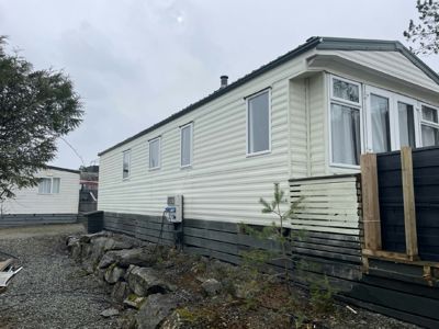 Fotografi av en hvit, paneltett hytte sett fra siden. Bildet viser fasaden, vinduer, tak, en del av grunnmuren og omgivelsene.