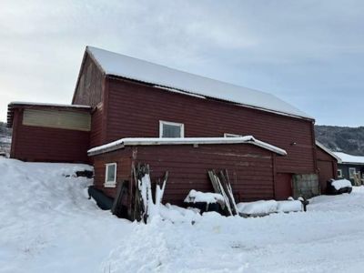 Fotografi av en rød, langhusformet bygning (sannsynligvis en uthus eller skur) sett fra siden. Bygningen har et skråtak med snø, vinduer og er omgitt av snø. I bakgrunnen ser man fjell.