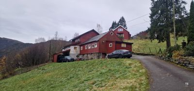 Fotografi av en rød, trebeslått bygning (sannsynligvis en gård eller hytte) sett fra en vei, med bil og omgivende natur.