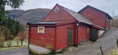 Fotografi av en rød, trepanelt bygning (sannsynligvis en uthus eller garasje) med en grønn dør, sett fra siden. Bakgrunnen viser skog og fjell.