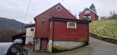 Fotografi av en rød, trepanelt bygning (sannsynligvis en uthus eller garasje) med en bil parkert foran og en annen bygning i bakgrunnen.