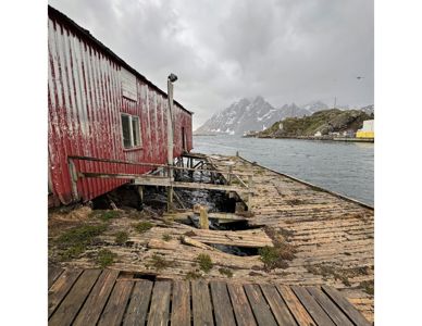 Fotografi av en rød, gammel bygning ved vannkanten med et utslitt tredekk. I bakgrunnen er det fjell og en by ved vannet.
