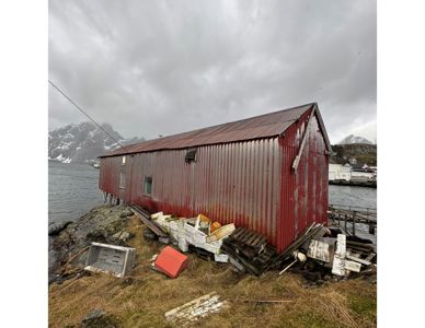 Fotografi av en rød, lang bygning (troll) på en klipperand ved vannet, med fjell i bakgrunnen.