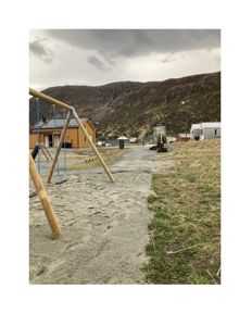 Fotografi av et utendørs område med en sandbunn, en gule bygning i bakgrunnen, en graver og en fjellside.