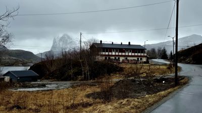 Fotografi av en rød og hvit bygning (sannsynligvis et hotell eller pensjonat) plassert på en høyde ved siden av en vei, med fjell i bakgrunnen.