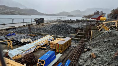 Fotografi av et byggeplass ved vannet med grunnarbeider i gang, inkludert armering og materialer.