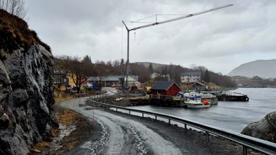 Fotografi av et byggeplass ved en kystvei med en stor kran, båter og boliger i bakgrunnen.