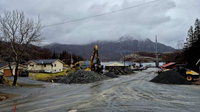 Fotografi av et bygge- eller anleggsområde med grusveier, maskiner, materialer og boliger i bakgrunnen.