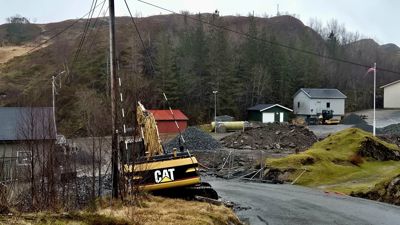 Fotografi av et byggeplassområde med grus, en gullgravemaskin og småbygninger.