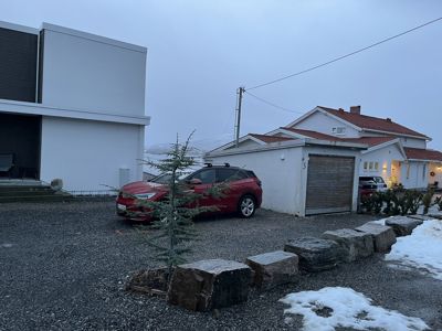 Fotografi av en moderne hvit bygning (venstre) og en garasje med en rød bil parkert foran. Bildet viser også nabolaget og terreng.